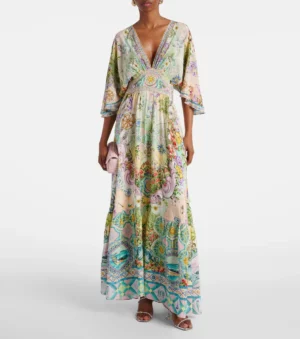 A person wearing a Camilla Printed Silk Crepe Maxi Dress with a V-neckline, three-quarter sleeves, and a colorful, intricate design stands holding a small pink clutch and wearing open-toe heeled sandals.