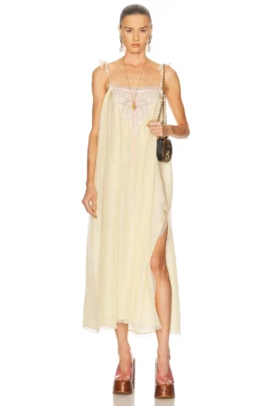 A person stands facing forward wearing a Chloe Lace-trimmed Silk Chiffon Midi Dress in light yellow with a side slit, platform brown shoes, a small shoulder bag, and gold jewelry against a plain white background.