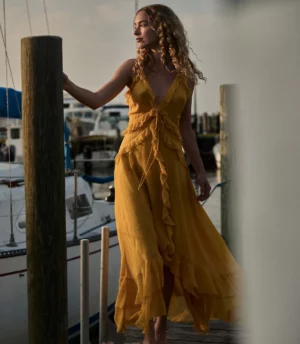 A woman in a sleeveless, yellow ruffled DÔEN Nevara Dress stands on a wooden dock next to a marina with boats in the background, looking to the side with one hand resting on a post.