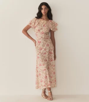 A woman stands against an off-white wall wearing the DÔEN Frieda Dress, a cream-colored, ankle-length floral piece with short ruffled sleeves. She has long dark hair and finishes the look with strappy gold sandals.