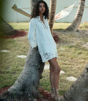 A woman with curly hair stands outdoors leaning against a palm tree, wearing a loose, white DÔEN Roselle Tunic with lace trim and metallic sandals. A hammock and grass are visible in the background.