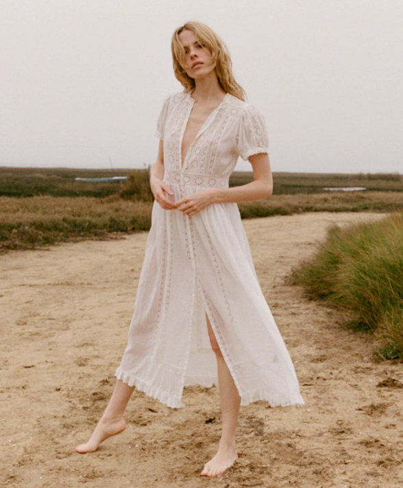 A barefoot person wearing a white, knee-length, lace dress stands on a dirt path in an open outdoor area with grass and overcast skies, channeling cool girl fashion.