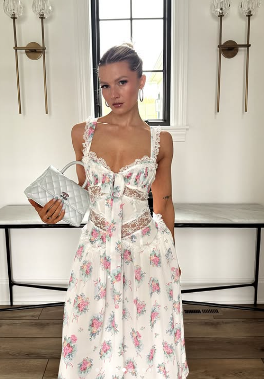 A woman stands indoors holding a small silver quilted handbag, wearing a white European Summer Dress with pink floral patterns and lace details. She poses in front of a window with two wall sconces on either side.