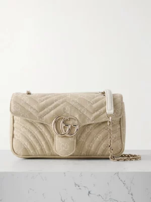 A beige Gucci Small GG Marmont Shoulder Bag with a gold "GG" logo on the front flap and a gold chain strap with a white leather section, displayed on a white marble surface against a light background.