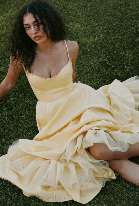 A person with curly hair wearing a butter yellow dress with spaghetti straps sits on green grass, looking toward the camera. The dress features a full skirt and visible tulle layers underneath.