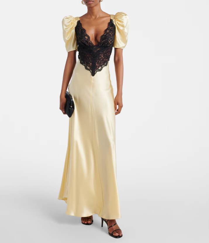 A person wearing a long butter yellow satin dress with puffed short sleeves and a deep black lace neckline, paired with black heeled sandals and a small black clutch.