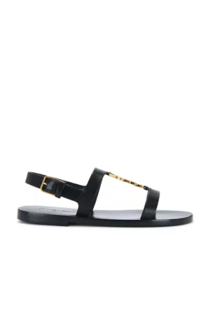 A flat black sandal inspired by the Saint Laurent Tribute Sandal, featuring a thin sole, open toe, T-strap with a gold accent, and adjustable ankle strap with a gold buckle. Photographed from the side on a white background.
