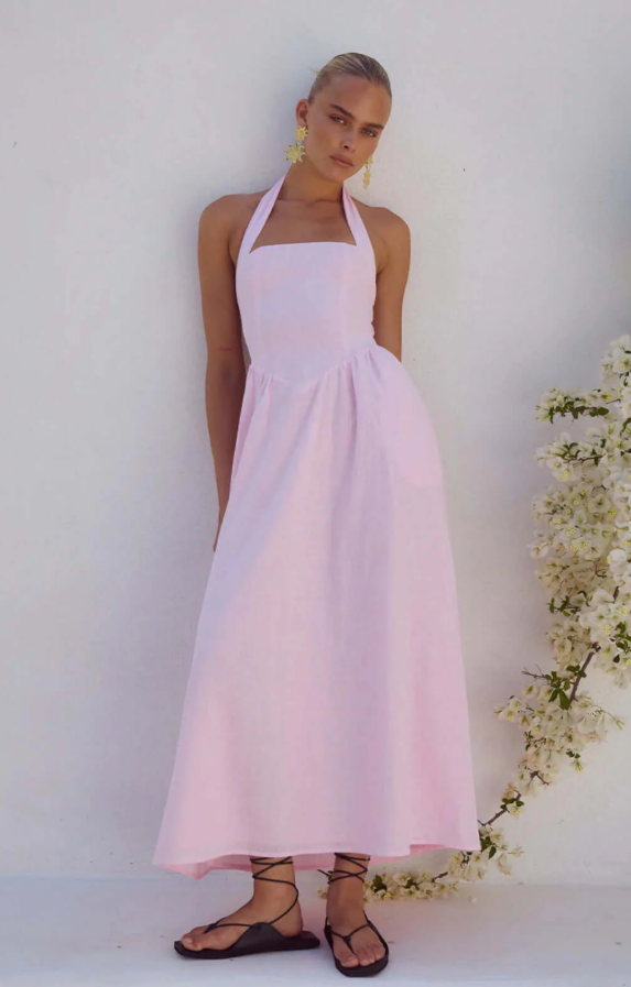 A person stands against a white wall wearing a light pink halter-neck dress with a fitted bodice and flowy skirt, channeling European Summer Dresses style, black lace-up sandals, yellow flower earrings, and branches with white flowers on the right.