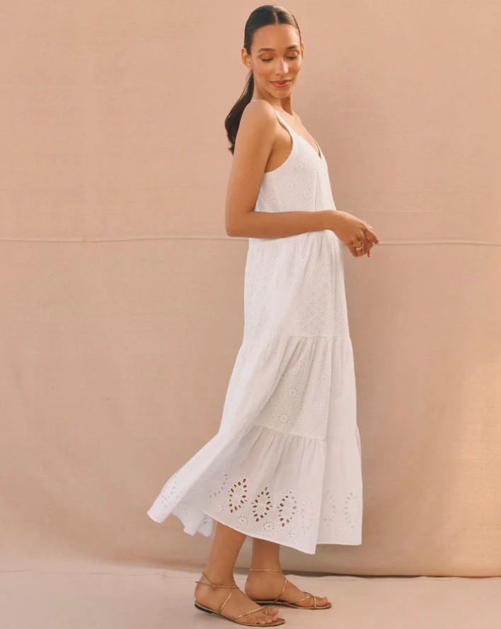 A woman stands sideways, wearing a sleeveless white eyelet maxi dress reminiscent of European Summer Dresses, with tiered layers and tan flat sandals. She has dark hair pulled back and poses in front of a plain beige backdrop.
