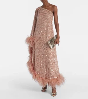 A person wears a one-shoulder, floor-length Ubud gown—a pink sequined gown with feather trim at the hem and sleeve. They hold a metallic clutch and wear metallic open-toe heels, standing against a plain white background.