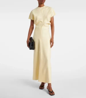 A person wearing a light yellow Toteme Gathered Maxi Dress with short sleeves, a high neckline, holding a black clutch bag and wearing black sandals stands against a plain white background.