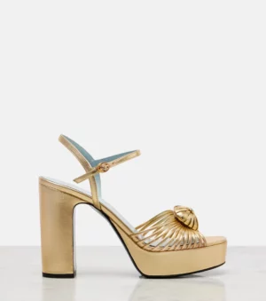 A gold high-heeled sandal with a thick platform sole, ankle strap with buckle, and shell-shaped detail on the front. This shiny Valentino Garavani Fawcette VLogo Signature 100 Leather Platform Sandal is displayed against a plain white background.