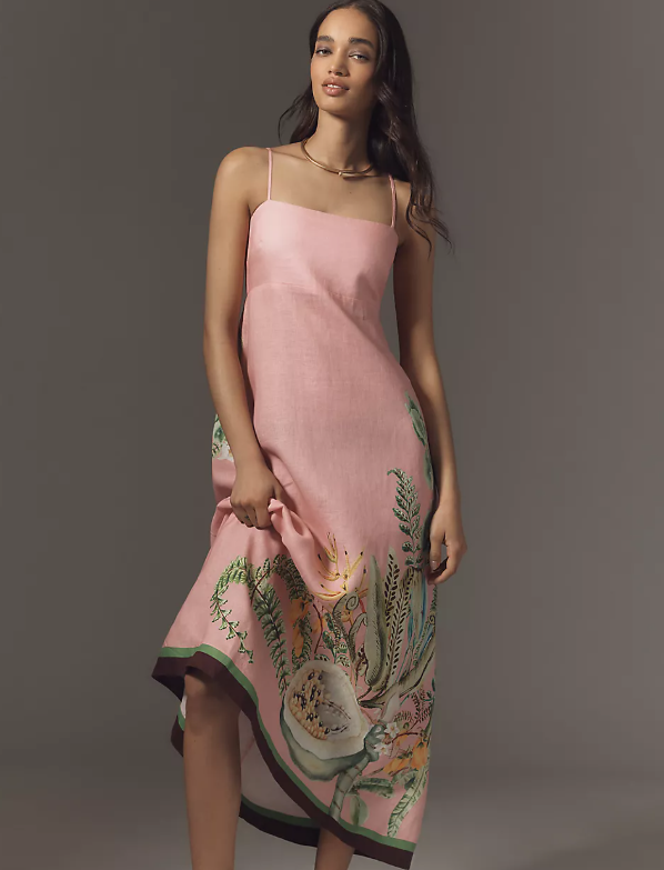 A woman stands against a plain background wearing a light pink summer dress with thin straps and a nature-inspired floral print along the lower skirt. She holds the side of her dress and looks forward with a neutral expression.