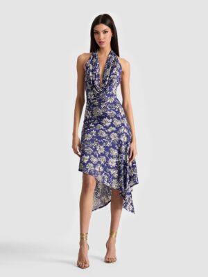 A woman stands against a plain background wearing the Alice + Olivia Dani Maxi Dress, a sleeveless blue and white floral piece with a draped neckline and asymmetrical hem. She has long dark hair and gold ankle-strap high-heeled sandals.