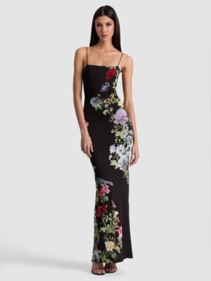 A woman stands against a plain background wearing the Alice + Olivia Dani Midi Dress, a long, fitted black dress with thin straps and a colorful floral pattern. She has straight dark hair and is wearing black heeled sandals.