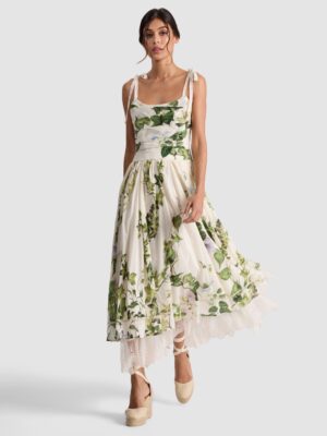A woman stands facing forward in a sleeveless, ankle-length white dress with green floral patterns reminiscent of the Alice + Olivia Chassidy Bell Sleeve Dress, featuring lace trim at the hem, bow straps, dark hair tied back, and beige platform shoes.