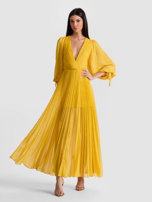 A woman stands against a plain background wearing the Alice + Olivia Oriana Drop Waist Midi Dress—a long, yellow, pleated dress featuring a deep V-neck and sheer, voluminous sleeves. She has straight dark hair and wears open-toed heels.