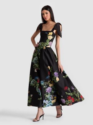 A woman stands in a sleeveless black Alice + Olivia Loryn Embroidered Shirt Dress with a colorful floral pattern, square neckline, and bow details on the shoulders. She pairs it with black strappy heels and has one hand on her hip.