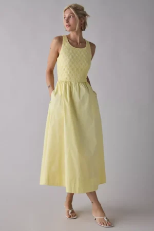 A person stands against a plain light background wearing a sleeveless, light yellow Bardot Mini Bow Dress with a fitted textured bodice and flared skirt, paired elegantly with white sandals.