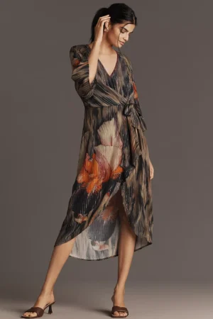 A woman stands against a plain background wearing the Bl-nk Ferlina Long-Sleeve Midi Shirt Dress. The dress features a brown and black pattern with large orange and beige floral prints, paired with brown heeled sandals.