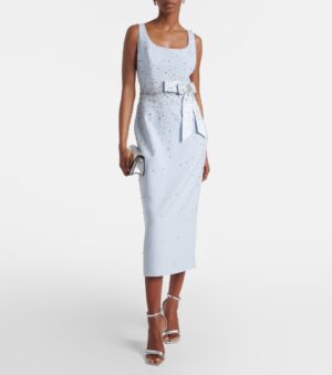 A woman wears a sleeveless, light blue Carolina Herrera Tweed Mini Dress adorned with small embellishments and a matching belt with a bow. She holds a silver clutch and wears strappy silver high-heeled sandals.