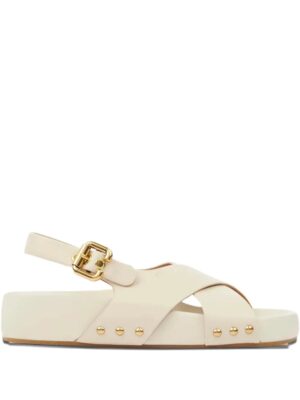 A beige sandal with a flat sole, crisscross straps, gold-tone buckle, and gold stud accents along the side. Inspired by the Chloé Kick Mule Sneaker, it features an ankle strap and open toe design for effortless style.