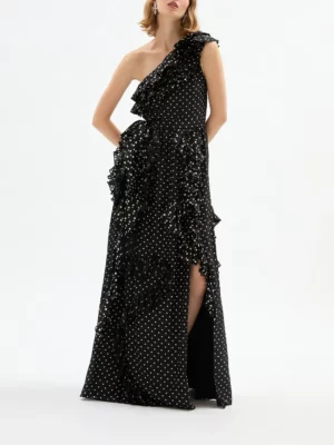 A woman wears the Delphine Angela Gown—a black, floor-length, one-shoulder dress with white polka dots and ruffled details. The gown features a side slit, and her arms are relaxed by her sides.