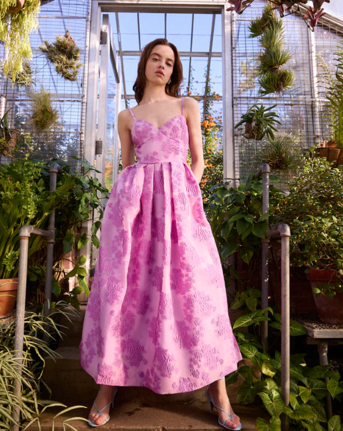 A person stands in a greenhouse wearing a long, pink, floral-patterned Hampton's wedding guest dress with thin straps. Green plants and hanging pots surround them as sunlight filters through the glass walls and ceiling.