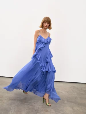 A person with a bob haircut wears a flowing, ruffled, blue Delphine Annette Midi Dress and green heeled sandals, walking indoors on a concrete floor against a plain white wall.