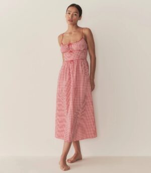 A person stands barefoot against a plain light-colored wall, wearing a sleeveless, ankle-length red and white gingham Dôen Angeline Dress with thin straps and a fitted bodice.