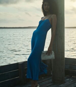 A person in a blue DÔEN Lorraine Dress stands on a wooden pier by the water, leaning against a post and holding a white textured clutch. The sky is cloudy and the water is calm in the background.
