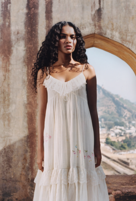 A woman with long curly hair stands in front of a weathered archway, wearing a white sleeveless flowy beach dress with delicate floral embroidery. A hilly landscape and buildings are visible in the background.