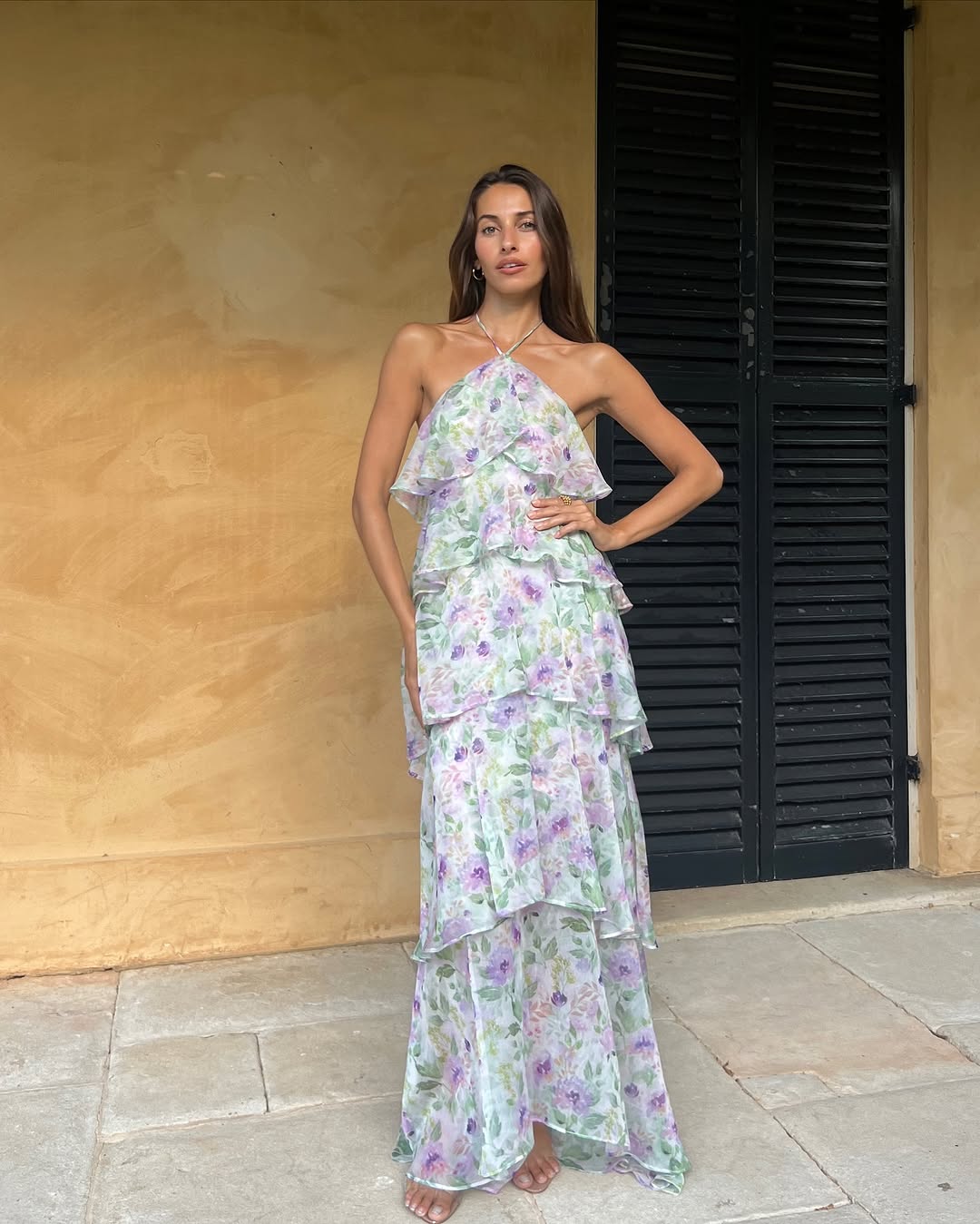 A woman stands barefoot on stone tiles in front of a tan wall and black shuttered door, wearing a ruffled, flowy lavender floral Hampton's wedding guest dress with a halter neckline. Her left hand rests on her waist as she looks at the camera.