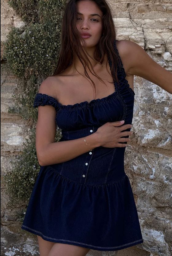 A woman with long brown hair wears a dark blue, off-the-shoulder, ruffled denim dress with front buttons—perfect inspiration for how to style your first date night dress—standing by a textured stone wall with greenery in the background.