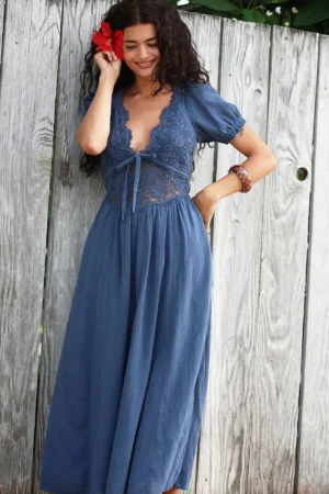 A woman with long curly hair stands by a weathered wooden fence, wearing the Free-Est Plenty of Love Maxi in blue. She holds a red flower near her hair, smiling slightly as lace detailing graces the bodice of her dress.