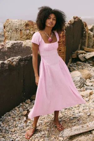 A woman stands on a rocky shoreline wearing a Free-Est Full Of Sunshine cotton-linen mini dress and sandals. She has curly hair, a large pendant necklace, and is framed by weathered stone and concrete walls in the background.