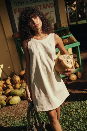 A person with curly hair in a Free-Est All You Need Is Lace Midi beige dress stands outdoors holding a coconut. Behind them are green crates filled with coconuts and a sign with partially visible text. Sunlight creates shadows on the scene.