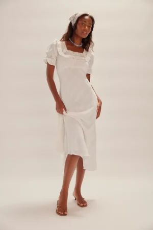 A woman stands against a plain background wearing the Free-Est Ciara Pucker Check Midi, a white, short-sleeved dress with lace details. She styles it with a printed headscarf, necklace, and heeled sandals, one hand resting on her hip.