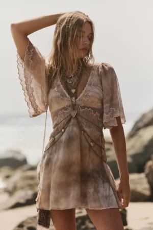 A woman stands on a rocky beach wearing the Free People Valeria Mini Dress, a light brown tie-dye short dress with lace trim and layered necklaces. She has wavy blonde hair and is looking down with one arm raised. The background is out of focus.