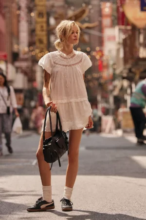 A person wearing a Free People Chance On Me Mini Dress, black sneakers, and white socks stands on a city street holding a black bag. The background features other people walking and blurred urban scenery.
