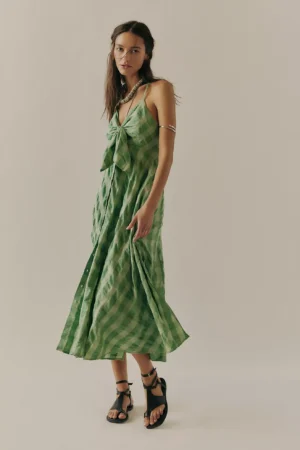 A woman stands against a plain background, wearing the Free People Winifred Mini Dress in green gingham with a front bow detail and black strappy sandals. She has long brown hair, glasses, and an arm cuff.