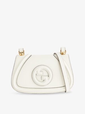 A small white Gucci GG Marmont Medium Woven Shoulder Bag with a curved shape, gold-tone hardware, and a prominent embossed logo on the front. The bag features a matching white shoulder strap draped across it.