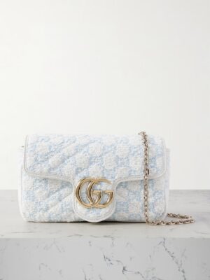 A light blue and white Gucci GG Marmont 2.0 Mini Leather-Trimmed Bouclé Shoulder Bag features a gold GG logo on the front flap and a gold chain strap, displayed on a marble surface against a plain background.
