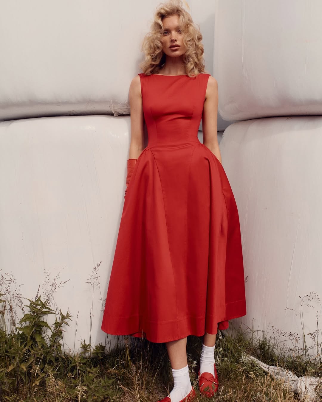 A woman with blonde, wavy hair stands in front of large white bales, showing how to style your first date night dress in a sleeveless, ankle-length red dress with matching shoes and white socks. Grass and weeds grow at her feet.