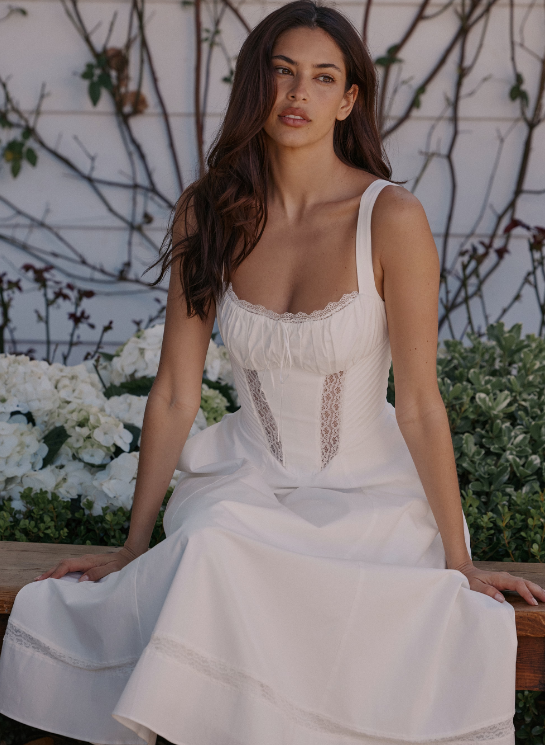 A woman with long brown hair sits on a wooden bench outdoors, wearing a sleeveless white dress with lace details—perfect for picnic dress ideas. Behind her are white flowers, green foliage, and a white wall draped with vines.