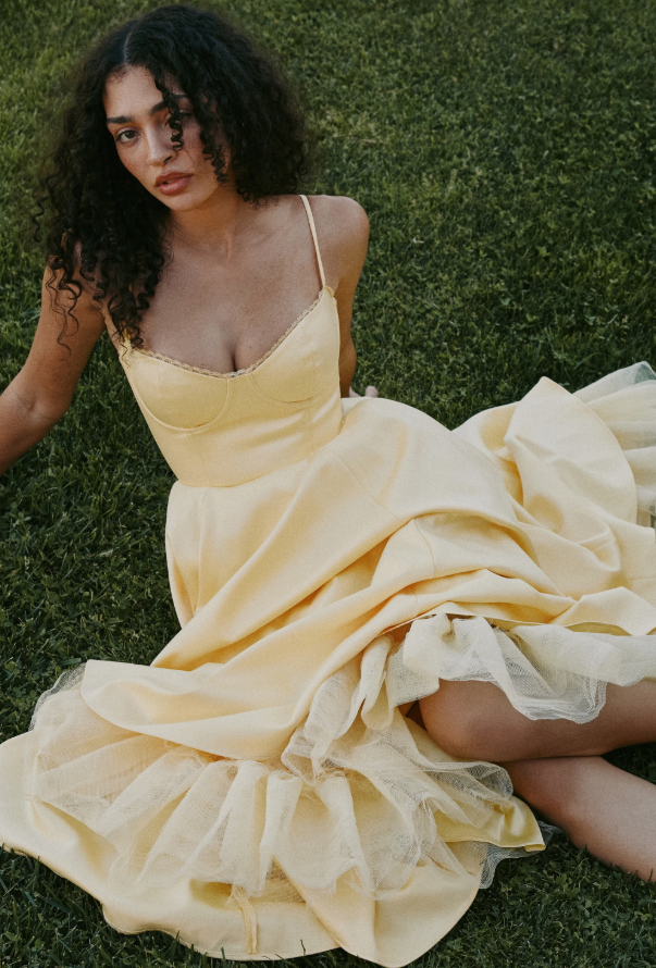 A person with curly hair wearing a light yellow Hampton's wedding guest dress sits on green grass, looking slightly upward. The dress features thin straps and a full, layered skirt.