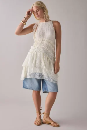 A woman stands against a plain background, wearing the Kimchi Blue Mackenzie Mesh Drop Waist Mini Dress layered over long, light blue denim shorts. She pairs it with tan sandals, gold bracelets, and hoop earrings, one hand touching her forehead.