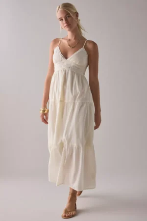 A woman stands against a plain background, wearing the Kimchi Blue Aria Strappy Tiered Ruffle Babydoll Mini Dress. She accessorizes her sleeveless white dress with gold jewelry and tan sandals for a simple yet elegant look.