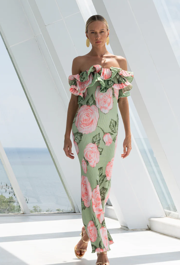 A woman stands indoors wearing a green off-the-shoulder, resort style dress with pink floral patterns and ruffled details. She has gold earrings and sandals, as sunlight streams through large windows behind her.