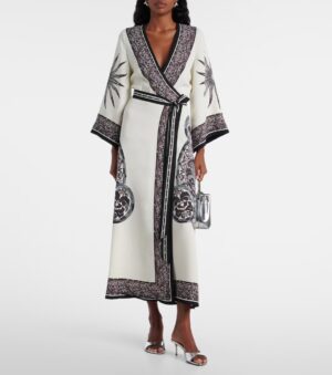 A woman wears a La DoubleJ Muumuu Printed Silk Twill Maxi Dress with black and silver ornate sun and floral motifs. She holds a silver handbag, pairs it with silver open-toe heels, and the dress features wide sleeves and a belt.